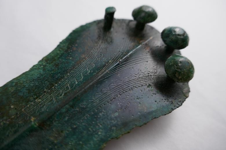 Ein Bild der Verzierungen des im Museum ausgestellten Dolches. Ein Teil eines Dolches mit grüner Patina und Verzierungen, die sich bogenförmig von der Mittelrippe zu den vier Nieten im Hintergrund ziehen