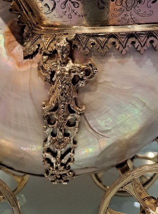 Zu sehen ist eine Nahaufnahme von einer der vier weiblichen Hermen, als silberne Verzierung des Nautilusgehäuses. Oben befindet sich der Kopf einer Frau, von dem aus sich Verzierungen aus silbernen Blumen  gerade nach unten um die Muschel schmiegen. Der Ornamentstrang verläuft zwischen zwei von vier Rädern auf denen das Gehäuse liegt. Alle vier Räder sind zu sehen, zwei im Vordergrund, zwei im Hintergrund. Aber alle nur teilweise, denn die Räder im Vordergrund sind durch die unteren Ecken des Fotos abgeschnitten und die Räder im Hintergrund verschwinden hinter dem Nautilusgehäuse. Die Räder sind golden. Einen Teil des Schiffskörpers mit schwarzen Ranken und silberner Spitze, die sich am Übergang von Schiff zu Nautilusgehäuse befindet, sieht man oben im Bild. Das Gehäuse glänzt perlmuttfarben , also rosa, weiß und türkis gemischt und hat schon ein paar kleine Risse. In der Knickstelle, wo sich das Gehäuse beginnt zu kringeln, ist es hellbraun. Auf dem Gehäuse sind außerdem kleine Schatten von den Rädern oder den Verzierungen, die leicht abstehen.