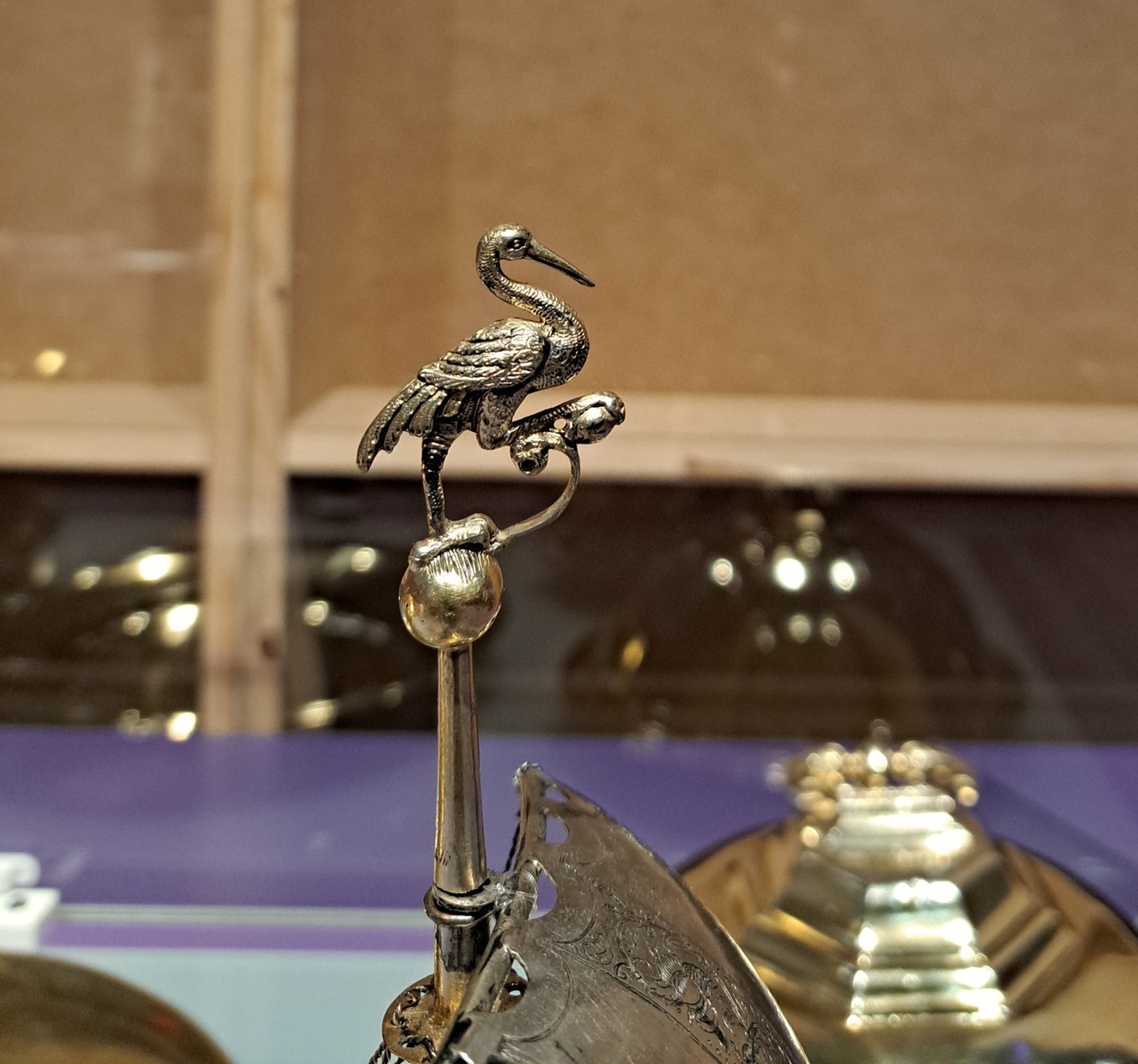 Zu sehen ist der Kranich des Tafelschiffs in Nahaufnahme. Der Kranich ist relativ mittig im Bild. Man sieht das Ende eines goldenen Mastes auf dem eine goldene Kugel sitzt. Auf dieser Kugel sitzt ein goldener Kranich. Der Kranich schaut nach rechts, ist also ebenfalls von der Seite zu sehen. Mit seiner linken Kralle steht er auf der Kugel und mit seiner rechten Kralle hält er einen goldenen Stein, welcher wiederum durch eine goldene Verbindung mit der Kugel verbunden ist. Der Hals des Kranichs ist s-förmig nach hinten gebogen, sodass der Schnabel nicht über den Körper hinausragt. Unten im Bild sieht man auch noch ein Ecke des silbernen Segels mit ein bisschen Verzierung. Im Hintergrund sieht man das Glas der Vitrine, denn es spiegeln sich die anderen Objekte darin. Links sieht man es nur als golden-weißes leuchten, aber rechts sieht man blass und dunkel eine goldene Glocke, die man über das Essen stülpen kann, damit es warm bleibt. Davon sieht man nicht nur die Spiegelung, sondern auch einen echten Teil hinter dem Tafelschiff. Hinter der Vitrine ist der Untergrund lila und die Wand besteht aus dünnen quadratischen Holzplatten, die mit dünnen hellbraunen Streben verbunden sind.
