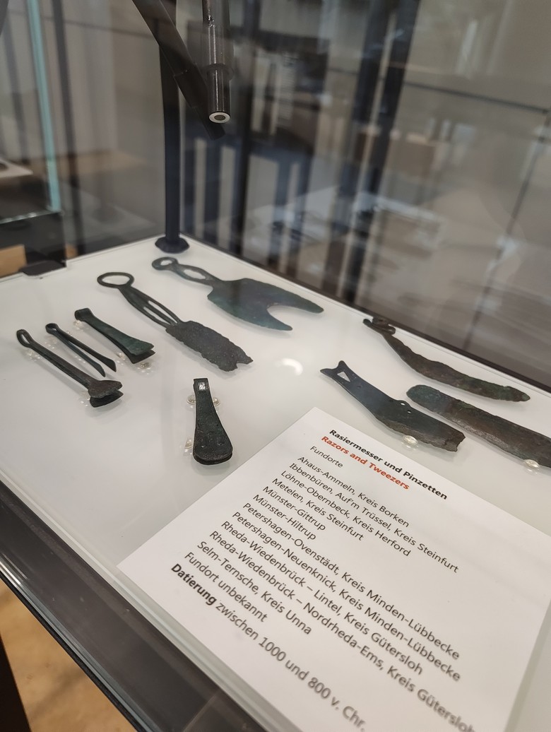 Foto von schräg rechts auf die Vitrine mit Fokus auf die Infotafel zu Fundorten und Datierung der Rasiermesser und Pinzetten. Oberhalb der Tafel, im rechten oberen Bildbereich, sind drei einschneidige Bronzerasiermesser zu sehen. Links daneben liegt ein sichelförmiges, doppelschneidiges Rasiermesser mit zweigeteiltem Griff und Endring. Darunter ist das für den Blogartikel ausgewählte zweischneidige Rasiermesser mit dreigeteiltem Griff zu sehen, sowie drei bronzene Pinzetten. Rechts daneben liegt eine weitere Pinzette schräg in der Vitrine.