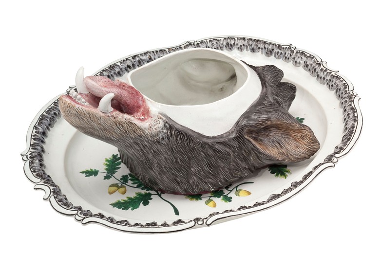 Das Bild zeigt eine Terrine in Form eines Eberkopfes wobei der Deckel abgenommen wurde. Die Terrine steht auf einem Teller, welcher eine Eichen Bemalung besitzt und eine Verzierung am Rand.