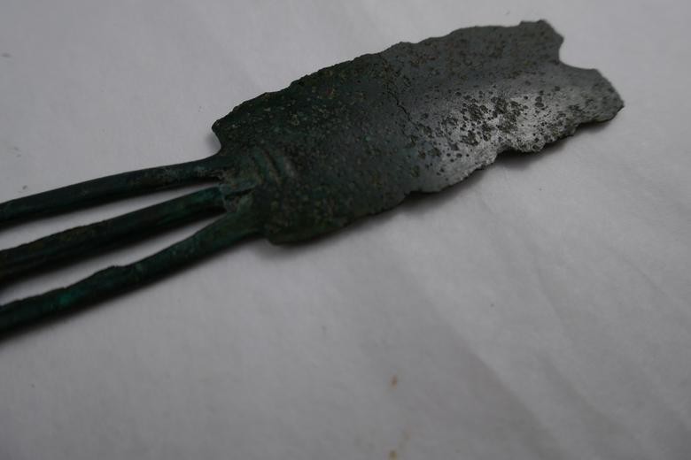 : Nahaufnahme der Klinge eines zweischneidigen Bronzerasiermessers aus der späten Bronzezeit. Im Bildvordergrund sind die drei durchbrochenen Griffstege zu erkennen, im Fokus liegt die stark korrodierte, langrechteckige Klinge mit unregelmäßigem Rand und einem V-förmigen Einschnitt an der oberen Kante. Die Oberfläche wirkt rau und dunkel oxidiert. Fotografiert auf hellem Untergrund, leicht seitlich von schräg unten.“