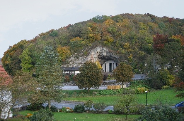 Ein Berg in dem die zugemauerte Balver Höhle zu sehen ist, auf dem Berg sind Bäume, vor dem Berg eine Straße und wiesen. Außerdem sind ein paar Häuser erkennbar.