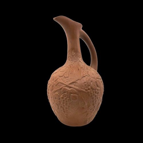Ein Tonkrug steht vor einem schwarzen Hintergrund. Der Krug ist nicht bemalt und hat einen langen dünnen Hals. Er hat einen Henkel.