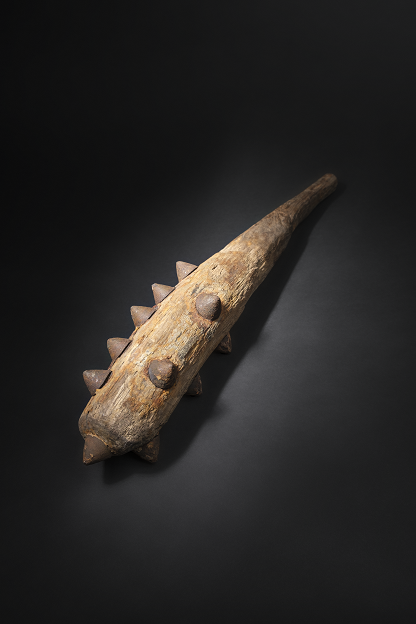 Keulenförmiges Holzstück mit Metallspitzen auf schwarzem Hintergrund.