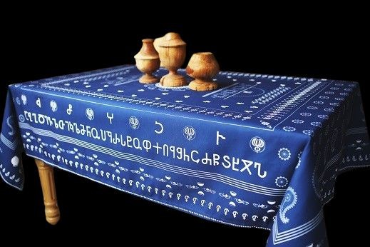Ein Tisch mit einer blauen Tischdecke steht vor einem schwarzen Hintergrund. Die Tischdecke ist mit weißen Zeichen, georgischen Alphabeten, verziert. Auf dem Tisch stehen drei Gefäße aus Holz.