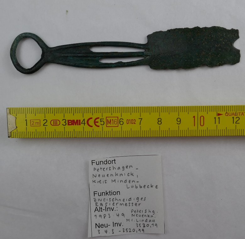 Foto eines zweischneidigen Bronzerasiermessers aus der späten Bronzezeit, liegend auf weißem Untergrund. Links befindet sich der ringförmige Griff mit drei durchbrochenen Stegen, rechts die stark korrodierte Klinge mit V-förmigem Einschnitt. Darunter liegt ein Maßband, das die Länge des Objekts mit etwa 12,5 cm angibt. Im unteren Bildbereich befindet sich ein handbeschrifteter Zettel mit Angaben zum Fundort (Petershagen-Neuenknick, Kreis Minden-Lübbecke), zur Funktion (zweischneidiges Rasiermesser) sowie zur alten Inventarnummer (1983.49) und zur neuen Inventarnummer (3.4.S – 3520,19).