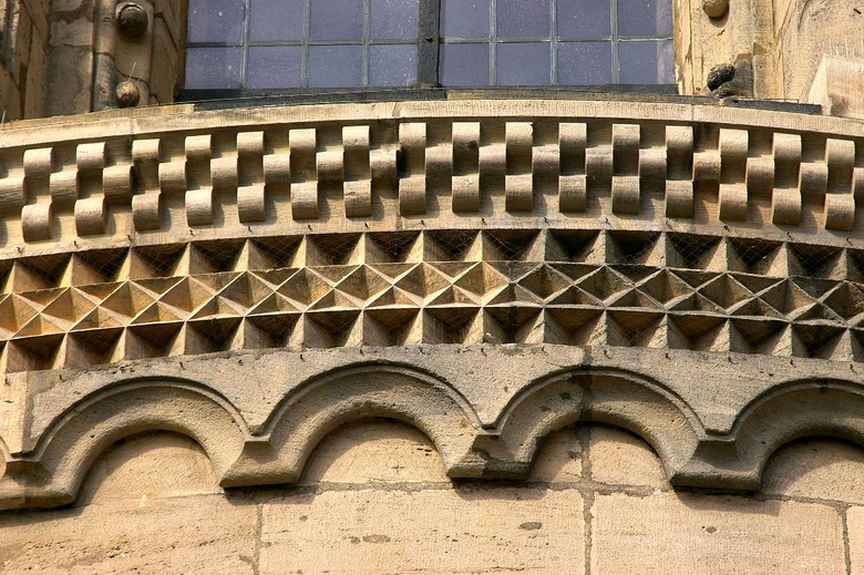 Das Bild zeigt einen Teil der Außenwand des Bamberger Doms. Dieser Bereich befindet sich unter einem Fenster und gehört zur sandsteinernen Außenwand, welche eine Verzierung aufweist, ein sogenanntes Fries. Dieser Fries verziert die Fassade und lässt sich in drei Teile unterteilen. Der oberste Teil hat ein sich wiederholendes Muster aus immer jeweils vier Quadraten, die sich an ihren Ecken berühren. Der mittlere Teil hat oben und unten ein Muster aus dreidimensionalen Dreiecken, die mit der Obersten Kannte aus der Wand heraus zeigend zu sehen sind, in der ist ein Muster aus sich an einander reihenden Rauten, welche mit ihren Spitzen oben und unten Dreiecke bilden. Der unterste Teil besteht aus dreidimensionalen Bögen.