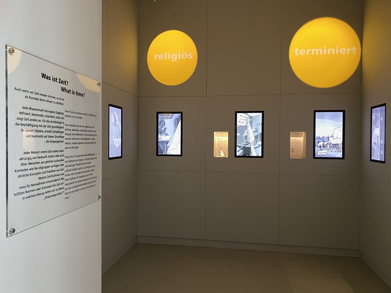 Blick in den Zeit-Kubus in der Dauerausstellung. Man sieht die Medieninstallationen und daneben die kleinen Vitrinen mit den Objekten. Die rechte Vitrine zeigt die Klappsonnenuhr.