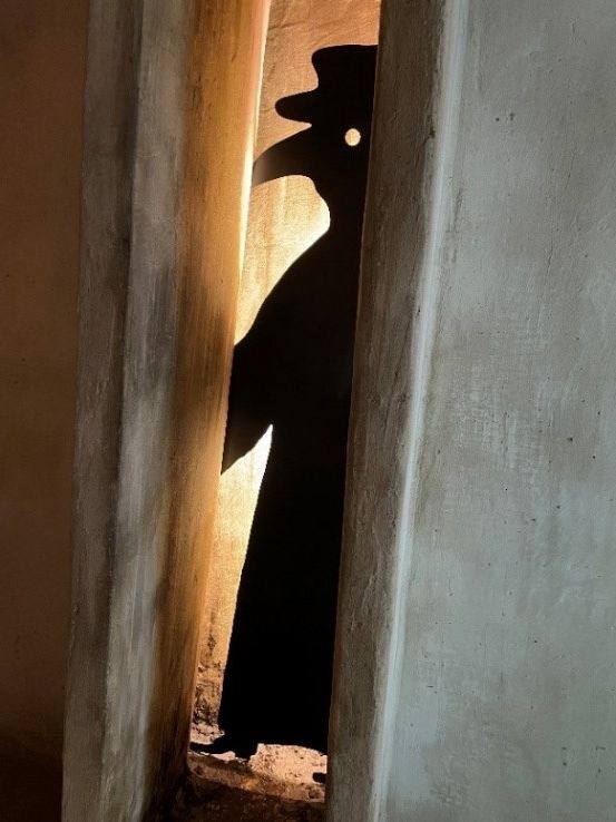 Auf dem Foto ist eine graue Betonwand zu sehen, in der ein Spalt ist. In diesem Spalt ist eine komplett schwarze Figur vom Seitenprofil zu sehen, die ein langes, weites Gewand, eine Schnabelmaske und einen Hut trägt. Das Licht kommt von hinten und gibt der Figur einen hellen Umriss.