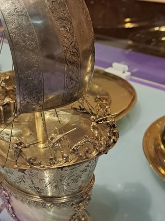 Zu sehen ist das Tafelschiff aus einer anderen Perspektive und in Nahaufnahme. Man sieht den ersten Teil des Schiffes schräg von vorne und zwar kommt es von links in das Bild. Ganz vorne am Bug sieht man Verzierungen, goldene Kringel und Bögen. Dort vorne steht ein Mann, der Trompete spielt. Weiter am Rand verteilt stehen pro Seite zwei Männer mit Hut und einem Gewehr. Das Gewehr halten sie zum Schießen bereit. Hinter diesen goldenen Figürchen ragen auf jeder Seite goldene Lanzen waagerecht in das Bild. Links, im Hintergrund des Schiffs, sieht man noch die Beine von der Person, die auf dem Thron sitzt. Der Boden des Schiffes leuchtet auch golden und man sieht den Anfang des goldenen Mastes, der in der Mitte des Schiffs steht und von dem silbernen Segel verdeckt wird,  dass nach rechts aufgebläht ist. Auf diesem Foto erkennt man auch die Verzierungen des Segels besser. Es ist in fünf dicke, senkrechte Streifen aufgeteilt, immer abwechselnd ein Streifen mit Verzierung, einer ohne. Die Verzierungen sind eingravierte Blumenranken. Auch hier sieht man goldenen, feine Ketten, die das Segel halten. Eine linke Kette ist direkt an das Segel gebunden und geht nach links aus dem Bild. Eine andere Kette hängt dort locker, ist aber an den Schiffsrand gebunden. Am Bug sind vier Ketten stramm am Schiffsrand angebunden. Immer zwei schließen den ersten Mann mit Gewehr auf beiden Seiten ein. Diese Ketten kommen unter dem Segel hervor. Der silberne Schiffskörper ist auch noch zu sehen, mit seinen schwarzen Blumenranken. Die silberne Spitze, die daran anschließt ist auch noch zu sehen, genauso wie zwei Ornamentstränge, vorne und links an der Seite. Diese sieht man allerdings nicht mehr genau. Eine Ecke des Nautilusgehäuses, auf dem das Tafelschiff steht, ist unten links rosa-schimmernd zu sehen. Die beiden anderen Ausstellungsstücke aus der Vitrine sieht man im Hintergrund. ein goldener Teller verschwindet hinter dem Tafelschiff, der andere goldene Teller wird rechts im Bild abgeschnit