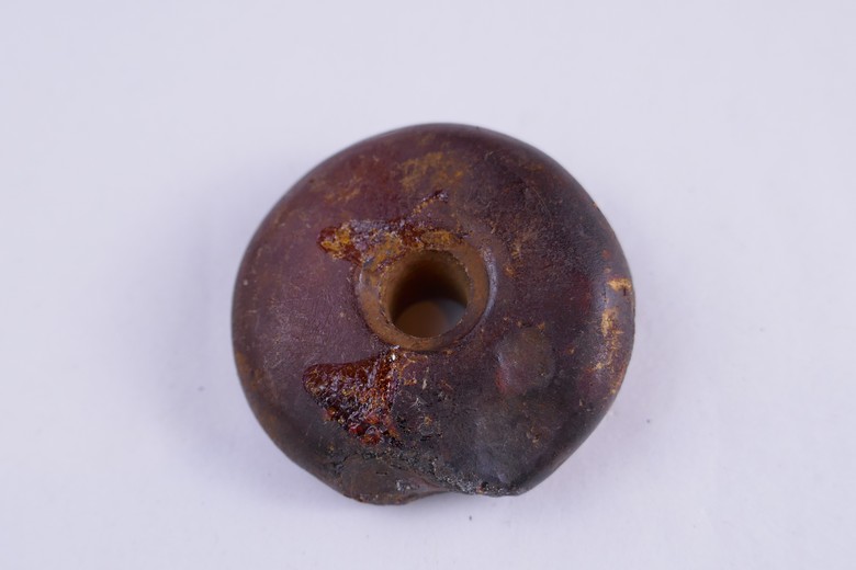 Auf dem Bild ist eine Bernsteinperle auf grauem Hintergrund zu sehen. Sie ist einer der kleinen mit einem 3-4cm Durchmesser, einem Loch zum Auffädeln in der Mitte und einigen Schrammen. Unten an der Perle und links und oben vom Loch sind kleine Stücke abgebrochen. Die Perle ist dunkelrot bis schwarz gefärbt und hat einige gelbe Einschlüsse.