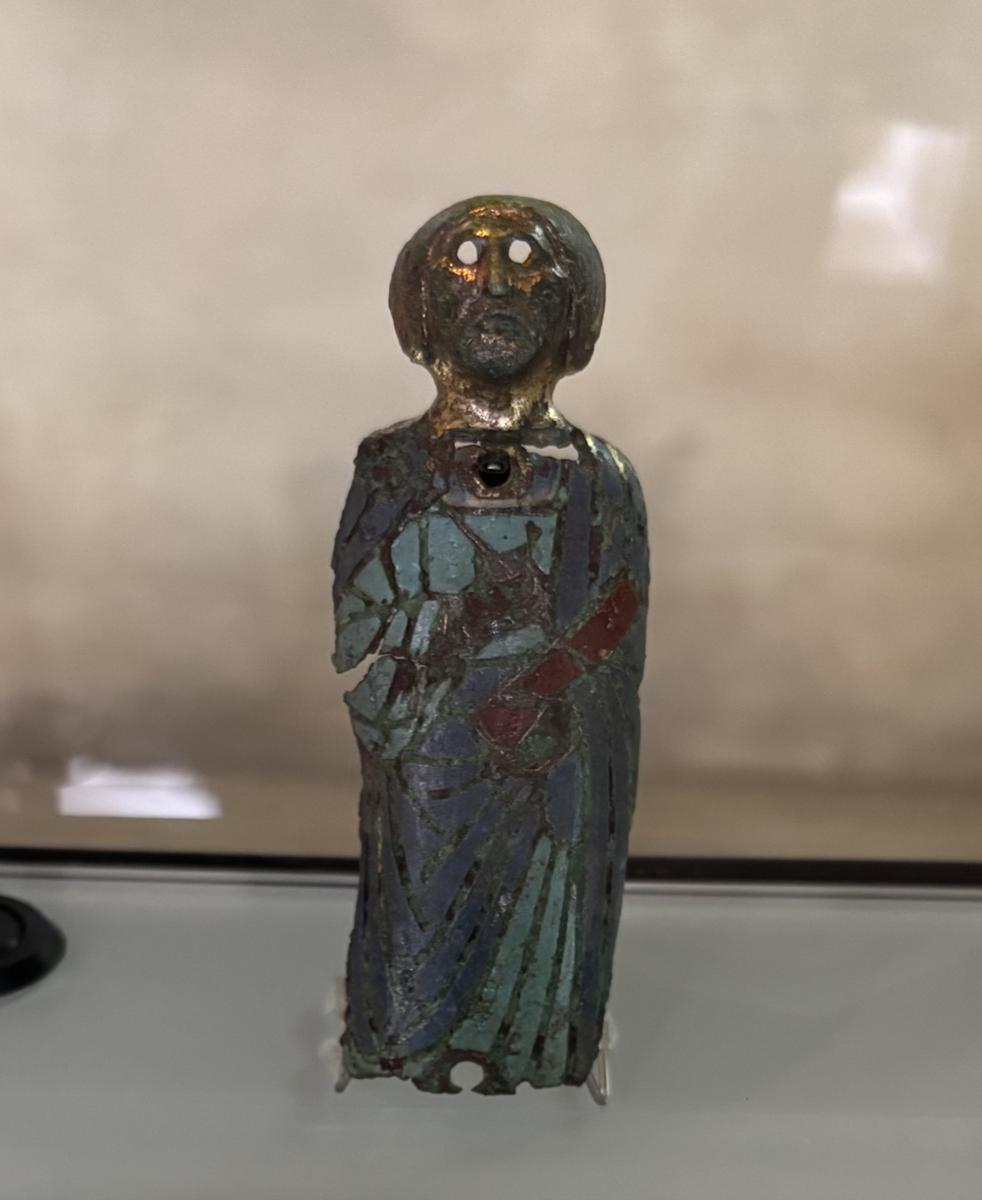 Kleine, etwa 10 Zentimeter große Figur des Apostels Petrus aus Buntmetall mit farbigen Emailleauflagen. Die stehende Figur trägt ein langes türkises Gewand und einen blauen Umhang. In ihrer rechten Hand hält sie einen Schlüssel, in ihrer linken Hand ein rotes Buch oder eine rote Schriftrolle. Die Augenhöhlen der Figur sind leer. Im Gesicht sind zusätzlich ein Bart sowie volles Haar dargestellt. Im Brustbereich befindet sich ein kleines Loch, eine weiteres befindet sich am unteren Rand der Figur. Beide dienten ursprünglich möglicherweise zur Befestigung auf dem Träger.