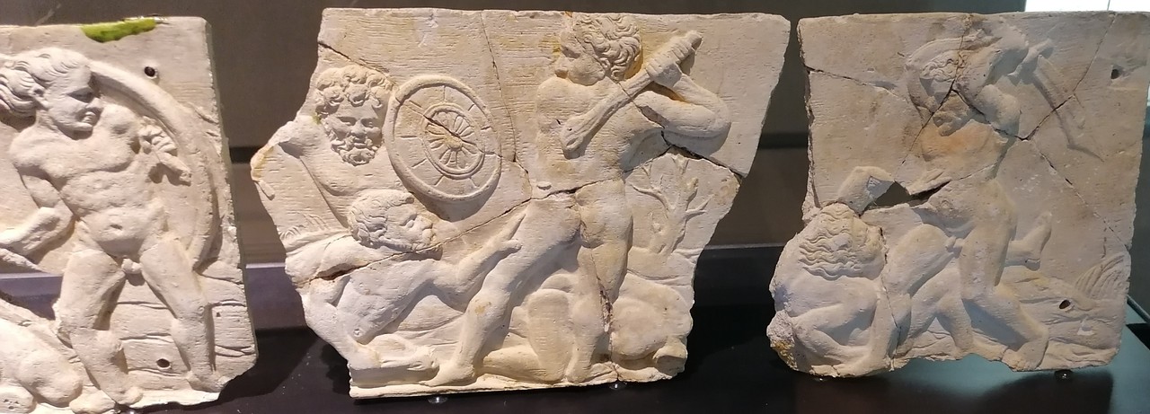 Das Bild zeigt eine Aufnahme aus einer Glasvitrine im LWL-Museum für Archäologie und Kultur in Herne. In dieser Vitrine ist ein Wandfries aus weißen Terrakottafliesen ausgestellt, welches Kampfszenen zwischen Hannibal und Scipio aus der Schlacht von Zama zeigt. Der Fries ist in drei Teile aufgeteilt und jeder Teil zeigt unterschiedliche Szenen. Ganz links ist ein Mann mit langen Haaren abgebildet, er hält etwas in den Händen, was nicht gut zu erkennen ist und steht vor einer Art Bogen, welcher auf einem Felsen steht, links neben ihm liegt etwas auf dem Boden, was ebenfalls ein Felsen sein könnte. Die Szene in der Mitte zeigt drei Männer, von denen zwei einen Bart tragen. Der Mann rechts hält eine Keule in der Hand, mit welcher er auf den Mann, der links zu seinen Füßen hockt, schlagen will. Dieser Mann berührt den Oberschenkel des Mannes mit der Keule und schaut zu ihm herauf. Links in der Ecke hinter den Beiden steht ein dritter Mann der einen Schild in der linken Hand hält und die Szene beobachtet. Im Hintergrund ist ein Strauch und ein Felsen zu sehen. Auf dem Friesstück ganz rechts wird eine weitere Kampfszene abgebildet, ein Mann liegt auf dem Boden und hält beschwichtigend eine Hand nach oben, während ein zweiter Mann, der über ihm steht und ein Schwert über dem Kopf hält, sich darauf vorbereitet den anderen Mann zu erschlagen. Im Hintergrund sind Felsen und in keiner Szene lässt sich erkennen welcher Mann welcher ist, da alle unbekleidet sind und es keine anderen Erkennungsmerkmale gibt.