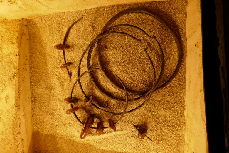Im zweiten Bild liegt der Bernsteinhort, in orangenem Licht getaucht, in seiner Vitrine. Er besteht aus fünf gerillten Ringen aus Bronze die aufeinanderliegend in Sand platziert sind. Die Ringe haben alle eine dunkelbraune und rostige Farbe und sind nach Größe und Dicke sortiert, vom kleinsten bis zum größten. Alle Ringe haben Rillen, aber diese werden dünner und enger mit jedem weiteren Ring. Auf dem ersten Ring sind 5 Bernsteinperlen, die wegen des Lichts karminrot erscheinen, die größte der Perlen ist in der Mitte und die kleineren sind in einem Paar um sie herum. Der zweite Ring ist in zwei Stücke geteilt. An dem oberen, längeren, Stück befinden sich zwei Perlen und an dem unteren Stück nur eine. Die Äußerste und letzte Perle ist auf dem dritten Ring aufgefädelt. Die zwei letzten Ringe haben keine Perlen.