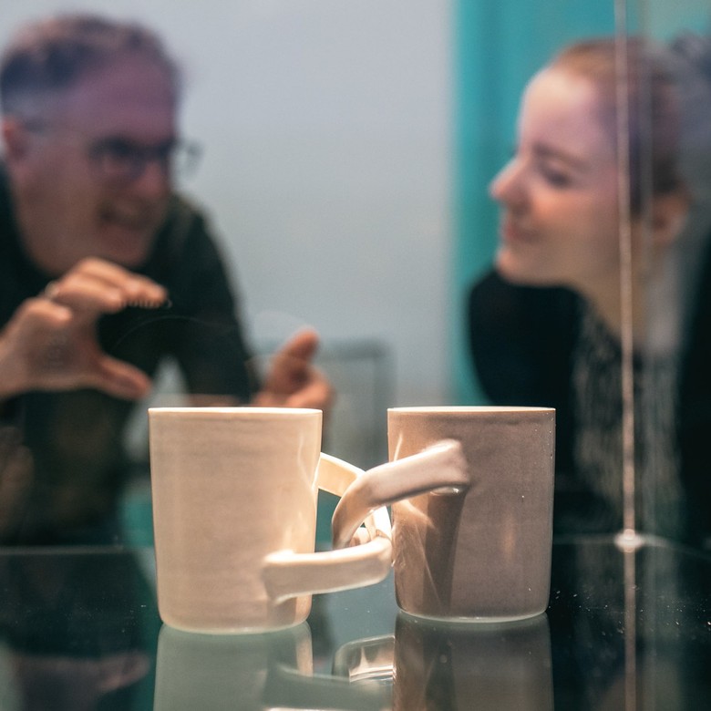 Im Vordergrund sieht man zwei Tassen, eine Graue und eine Weiße, die am Henkel miteinander verbunden sind. Im Hintergrund sieht man verschwommen einen Mann und eine Frau, die sich über diese Tassen unterhalten.