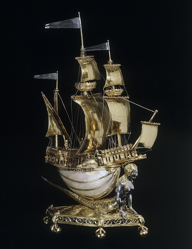 Zu sehen ist das Burghley-Nef in der Mitte des Fotos und der Hintergrund ist schwarz. Es ist von schräg vorne zu sehen, sodass die vordere Spitze nach rechts zeigt. Außerdem reicht es von der unteren Bildkante bis zur oberen. Dieses Tafelschiff sieht ganz anders aus als das Tafelschiff, um das es geht. Es ist komplett golden bis auf einzelne Teile. Das Deck läuft vorne und hinten spitz zu und diese Spitzen sind eingezäunt, sodass am Bug und am Heck Dreiecke entstehen. Ungefähr auf der Mitte des Zauns verläuft ein Querbalken, der die Streben verbindet. Überall schauen, mit ein bisschen Abstand, kleine Kanonenrohre zwischen den Zaunstreben hindurch.  Vorne an der Spitze vom Bug erkennt man einen Kopf, der vielleicht einen Drachenkopf darstellen soll. Man sieht vier Figürchen auf dem Schiff. Zwei Männer, je einer an der Ecke des Dreiecks auf der Seite, die dem Betrachter zugewandt ist. Dann ein Soldat mit einer Lanze in der Mitte des hinteren Dreiecks und ein Mann, halb verdeckt durch ein Segel, an der anderen Ecke des Dreiecks, also im Hintergrund. An der Spitze des Hecks steht ein Mast, an dem ein dreieckiges Segel schräg hängt. Auf dem Mast steht eine dreieckige silberne Fahne. Die Spitze zeigt nach links. In der Mitte des Schiffes ragt ein großer Mast in die Höhe. Er ist größer als der hintere Mast. Als erstes ist an diesem Mast das größte Segel befestigt, dass von der Mitte des Mastes fast bis zum Schiffsboden reicht. Es ist viereckig und nach rechts aufgebläht. Über dem Segel ist ein runder Ausguck rund um den Mast befestigt. Er sieht etwas aus wie eine Krone. Über diesem Ausguck ist ein sehr kleines viereckiges Segel befestigt und ganz oben auf dem Mast steht wieder dieselbe Fahne, wie auf dem anderen Mast. Ein dritter Mast steht im Zaundreieck am Bug. Er ist etwas kleiner als der hohe Mast, aber noch deutlich höher als der hintere Mast. Der Mast am Bug sieht so aus wie der hohe Mast, nur dass das unterste Segel viel kleiner ist, also nicht bis zum Boden reicht.