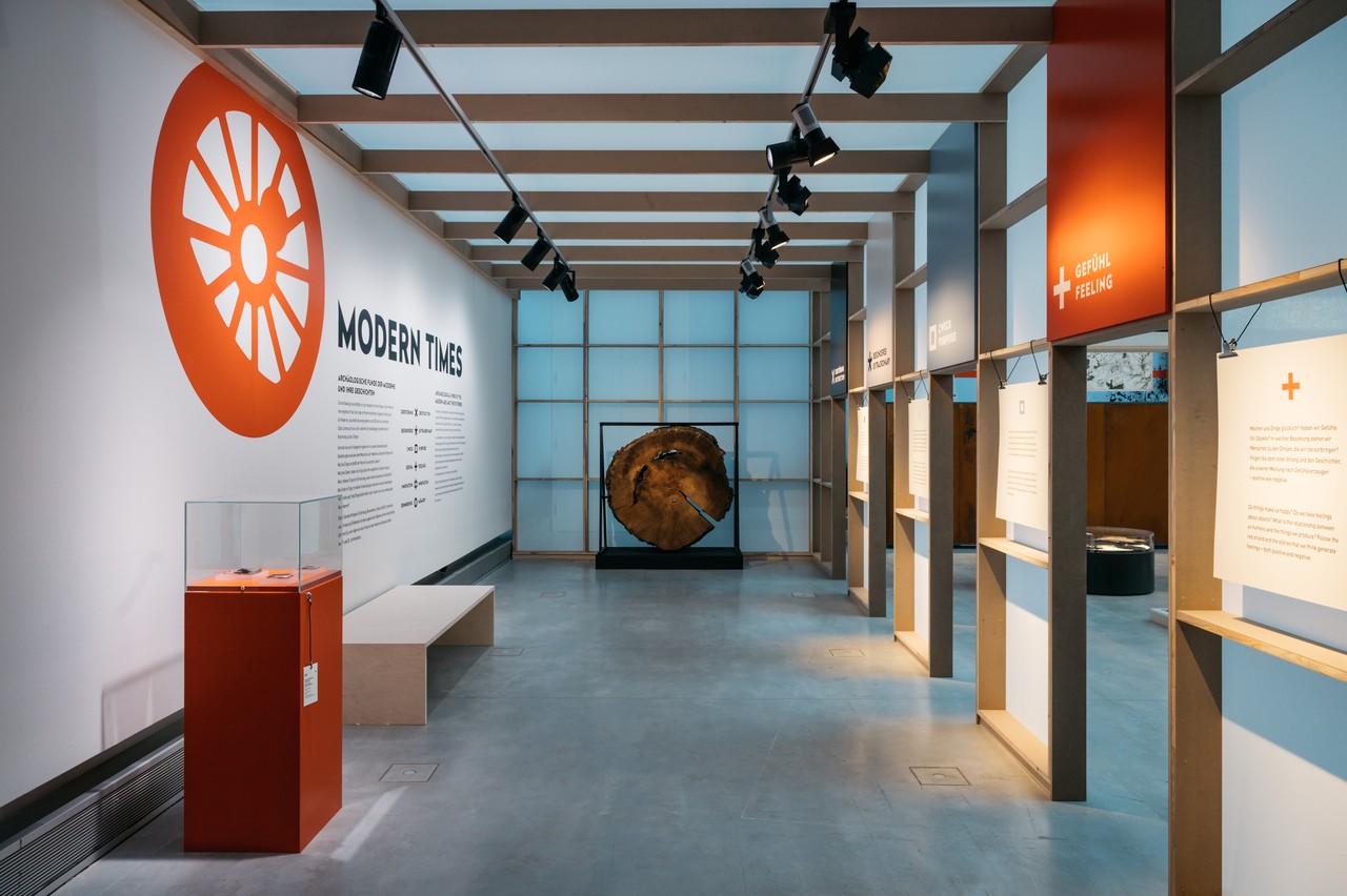 Ein Blick in den Eingangsbereich der Ausstellung. Links eine Wand mit Schrift und eine rote Vitrine mit Objekten. Am Ende steht eine Baumscheibe als Objekt.