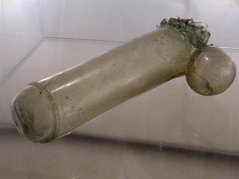 Fotografie eines Glasgefäßes in Form eines Phallus in einer Vitrine.