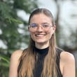 Portrait von Tabea Doll. Die Haare sind halb hochgesteckt und sie trägt eine runde Brille und ein schwarzes Top. Sie grinst freundlich in die Kamera.