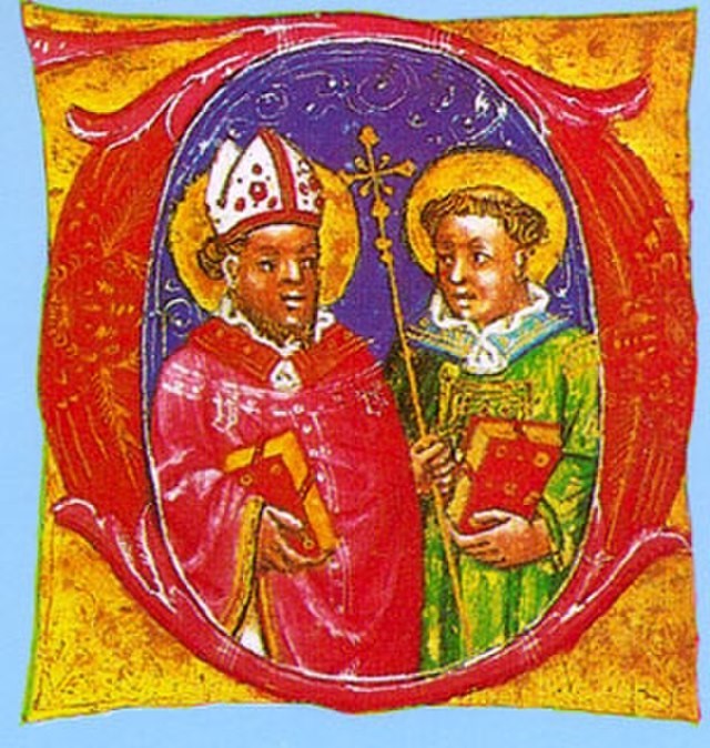 Das Bild ist ein Kunstwerk. Es zeigt den Bischof Sankt Hermagoras von Aquileia und seinen Diakon Sankt Ermagora umrandet von einem roten Kreis und einem goldenen quadratischen Rahmen. Der Bischof trägt ein rotes Gewand mit Musterungen und goldenen Streifen an den Rändern. In seiner rechten Hand hält er ein rotes Buch mit goldenen Seiten, vermutlich die Bibel. Auf seinem Kopf trägt er eine weiße Bischofsmütze mit roten Mustern. Er besitzt einen goldenen Heiligenschein. Sein Diakon trägt ein grünes Gewand mit goldenen Mustern und einem türkisen Kragen, der auch golden gemustert ist. Er hält in der rechten einen goldenen Stab mit einem prunkvollen Kreuz und in der linken das gleiche Buch wie der Bischof. Auch er besitzt einen goldenen Heiligenschein. Die beiden kirchlichen Würdenträger vermitteln durch ihre edle Kleidung und die goldenen Heiligenscheine Reichtum und Macht.