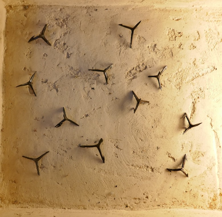 Eine Perspektive von Oben auf die, in den Boden eingelassene Vitrine in der Dauerausstellung des LWL-Museums für Archäologie und Kultur in Herne, welche die Krähenfüße beinhaltet. Elf Krähenfüße sind auf einer Fläche, die etwa die Größe einer Bierkiste hat, verteilt. Der Blickwinkel ist genau von Oben auf die Vitrine und deshalb lassen sich kaum die Spitzen erkennen, welche nach oben weisen. Manche der Krähenfüße weisen sichtbare Wiederhaken auf. Die jeweiligen Spitzen sind in etwa vier Zentimeter lang. Alle Krähenfüße in der Vitrine sind rostfarben. Sie liegen auf einem Untergrund, welcher nach glattgeputztem Sand oder sogar Sandstein aussieht.