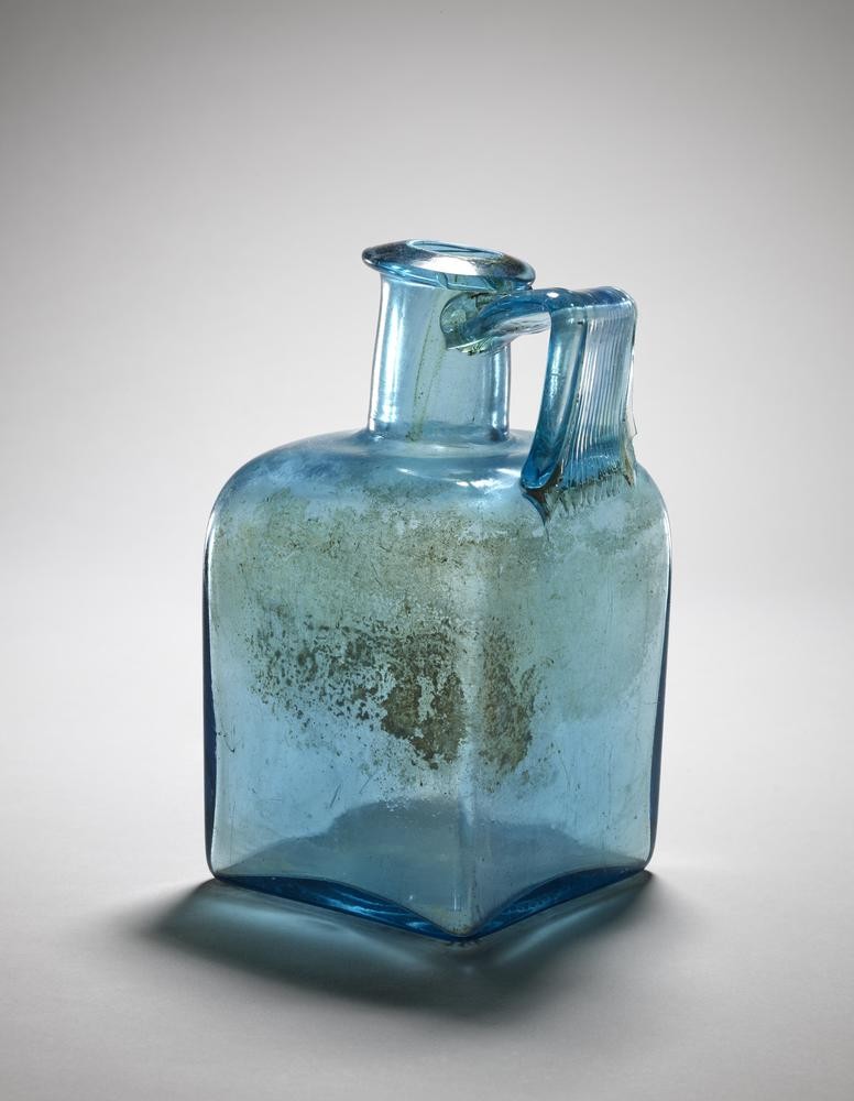 Zu sehen ist eine typische römische Vierkantflasche auf weißem Hintergrund. Diese Flasche ist durchsichtig, aber bläulich schimmernd.Sie besitzt eine rechteckige Grundfläche. Sie ist relativ klein, dafür breit und hat einen kurzen Hals mit Henkel. Der Bauch sieht verschmutzt aus.