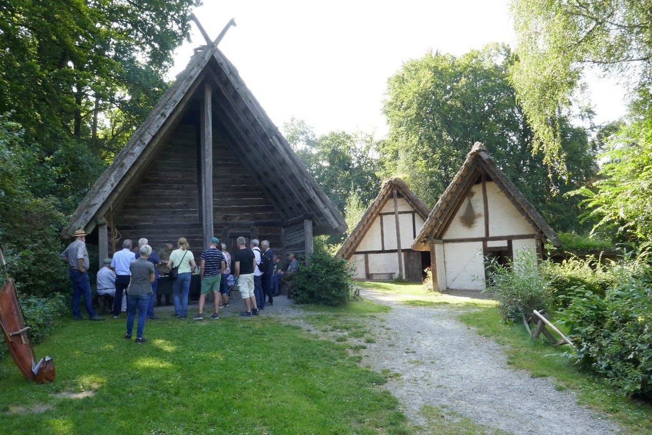Eine Menschengruppe sammelt sich im Schatten vor einem der drei abgebildeten Hütten des Freilichtmuseums Oerlinghausen, und scheint einer Führung zuzuhören.