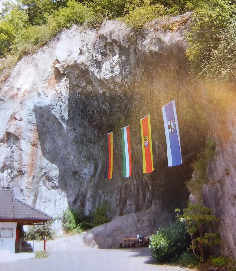 Hier wird die Balver Höhle von vorne rechts gezeigt. Sie hat eine riesige Öffnung. Vor der Höhle links unten im Bild ist ein Teil eines Hauses zu sehen, das deutlich kleiner ist als die Höhlenöffnung. Um die Höhle herum wachsen verschiedene Büsche und Bäume. Nur der gepflasterte Gehweg und das Höhleninnere sind unbewachsen. Oben am Eingang der Höhle sind 4 große Fahnen an einem waagerechten Seil angebracht. Die Flaggen sind von links nach rechts: die Deutschlandflagge (schwarz, rot und gelb), die NRW-Flagge (grün, weiß und rot), die Flagge des Märkischen Kreises (gelb und rot, mit einem Wappen in gelb, rot, weiß und schwarz) und die Flagge der Stadt Balve (weiß und blau, mit einem Wappen in schwarz, weiß und blau).