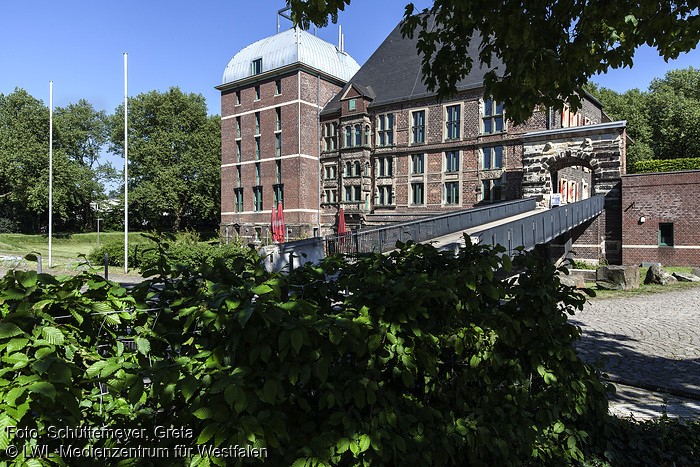 Das Bild zeigt Schloss Horst von außen. Vorne im Bild sieht man grüne Blätter von den umliegenden Büschen und Bäumen. Der bogenförmige verzierte Steineingang des Schlosses ist durch eine Art Brücke, welche über einen grauen Kopfsteinpflasterweg verläuft, zu erreichen. Der Weg führt zum Park, der mit Büschen, Bäumen und Rasen bepflanzt ist und das Schloss umgibt. Im Park stehen außerdem zwei Fahnenmaste, ein Mülleimer und einige Steine. rechts neben dem Eingang befindet sich ein eckiges Haus aus roten Ziegeln mit einem grünen Fensterrahmen, unter dem Fenster verläuft ein weißer Streifen. Links neben dem Eingang befindet sich das eigentliche Schloss, das aus zwei sichtbaren Gebäudeteilen besteht. Vor dem Schlossgebäude befinden sich noch zwei rote Sonnenschirme, die trotz des sichtlich sonnigen Wetters geschlossen sind. Der sich näher am Eingang befindende größere Gebäudeteil besteht aus roten Ziegeln, vierundzwanzig von vorne sichtbaren Fenstern mit grünem Fensterrahmen, von denen neun Rundbogenfenster sind, und einem schwarzen Dach, welches die Form einer Pyramide mit rechteckiger Grundfläche hat. Von diesem Gebäudeteil ist auch die rechte Seite sichtbar, die sich dort befindenden Fenster haben rot/weiße Fensterläden. Der weiter linksstehende Gebäudeteil ist schmaler als der andere, ebenfalls aus Ziegelsteinen und besitzt sechstzehn Fenster mit grünem Rahmen unter denen ein weißer Streifen verläuft. Das Dach ist eine aus grünem Kupfer bestehende Kuppel mit rechteckiger Grundform. Auf dem Dach befinden sich zwei Metallstäbe und ein nicht zu erkennendes Metallkonstrukt.