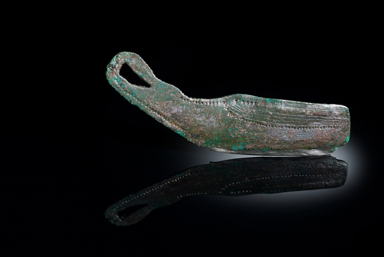Das beschriebene bronzezeitliche Rasiermesser mit der Schiffsdarstellung. Am Rand sind Punkte, im rechten oberen Quadranten sind sechs horizontale Linien eingepunzt, das Messer ist grün-bräunlich mit grünen Oxidierungen, der Hintergrund des Bildes ist dunkel, das Messer spiegelt sich leicht im Boden