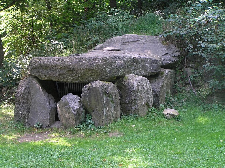Auf dem Bild sieht man sechs hochkant stehenden Steine, die in einem leichten Rechteck gestellt sind. Darauf liegt ein großer Stein, der als Deckel dient. Die Steinkiste steht draußen in einer Grünfläche mit Gebüschen und Bäumen.