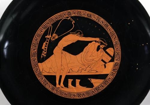 Zu sehen ist der Boden der Innenseite der Kylix. Hier liegt ein älterer, bärtiger, bekleideter Teilnehmer seitlich und aufrecht auf einer Kline und führt einen Finger zum Mund. Ein langhaariger, jüngerer, nackter Mann steht neben der Kline und hält ihm den Kopf. Unter oder neben der Kline findet sich ein Paar Schuhe und eine Schüssel, in die sich der ältere Mann zu übergeben versucht. Über ihren Köpfen schweben eine Leier und ein Flötenfutteral. Eine runde verzierte Borde rahmt die Szene ein. Die Szene nimmt etwa die Hälfte der Fläche der Unterseite der Kylix ein.