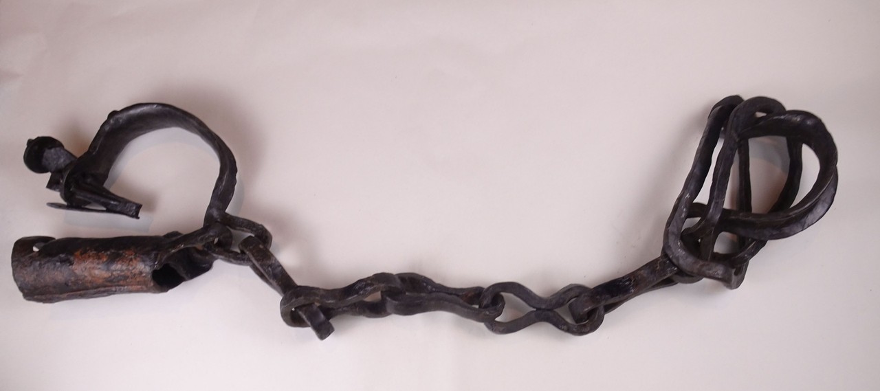 Es ist eine Fußfessel auf einem weißen Untergrund zu sehen. Diese Fußfessel hat auf der linken Seite etwas Rost und sie besteht aus Schmiedeeisen und Stahl. Die zwei großen Ringen auf der rechten und linken Seite sind mit fünf kleineren Ringen in der Mitte miteinander verbunden.