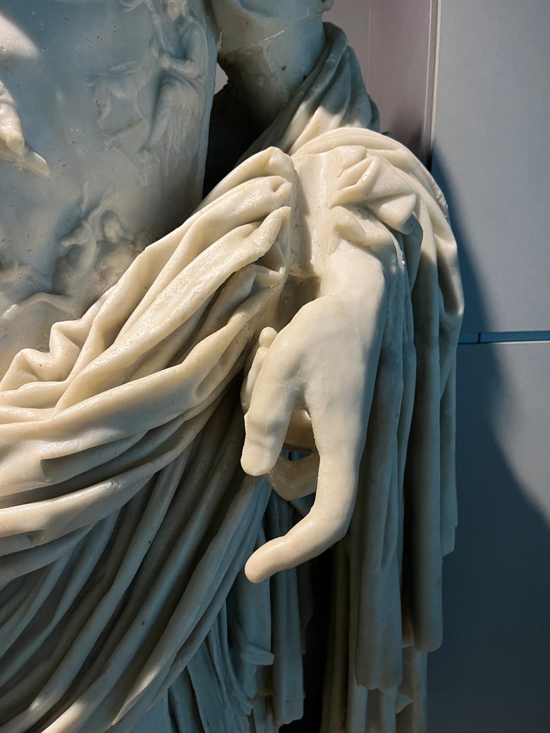 Das Bild ist eine Nahaufnahme von Augustus linker Hand, welche etwas geöffnet ist und leicht nach unten zeigt. Die Finger sind entspannt und leicht gekrümmt. Um den Arm und über der Hand häng ein Gewand in zahlreichen, kunstvoll übereinanderliegende Falten nach unten. Im Gewand, knapp über dem Handgelenk, ist eine schmale, runde, lange und glatte Vertiefung zu erkennen.