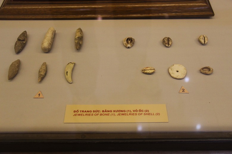 Das Bild zeigt eine Vitrine mit archäologischen Artefakten. Diese Artefakte sind als Schmuckstücke klassifiziert und bestehen aus Knochen und Muscheln. Links sind sechs längliche, unregelmäßig geformte Objekte zu sehen, die als Schmuckstücke aus Knochen gekennzeichnet sind. Diese Knochenartefakte haben unter-schiedliche Formen und Größen, einige sind spitz zulaufend, andere sind zylindrisch oder gebogen. Rechts befinden sich sieben kleine Objekte, die als Schmuckstücke aus Muscheln gekennzeichnet sind. Diese Muschelstücke variieren ebenfalls in ihrer Form, einige sehen aus wie kleine Muschelschalen, andere haben runde oder ovale Formen mit einem Loch in der Mitte. Unter den Objekten befindet sich ein Schild mit der Aufschrift: „Đồ Trang Sức: Bằng Xương (1), Vỏ Ốc (2) / Je-welries of Bone (1), Jewelries of Shell (2).“