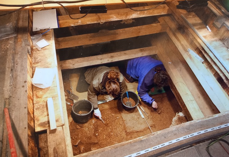 Auf dem Bild ist eine Grabung zu sehen. Zwei Frauen graben in einem quadratischen Loch. Die Wände werden von Holzbalken von allen 4 Seiten gehalten und stabilisiert. Beide Frauen knien mit dem Kopf in Richtung der Kamera und schauen dabei nach unten, somit sieht man von ihnen nur die Haare und nicht das Gesicht. Sie graben mit kleinen Werkzeugen in der Erde. Neben beiden Frauen steht ein Eimer mit etwa Erde darin.