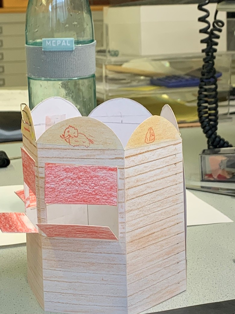Sechseckiges Papiermodell der Buddel Bude, angemalt mit Buntstiften, verziert mit Mammut-Abbildungen. Das Model hat drei aufklappbare Fenster und ist oben offen.