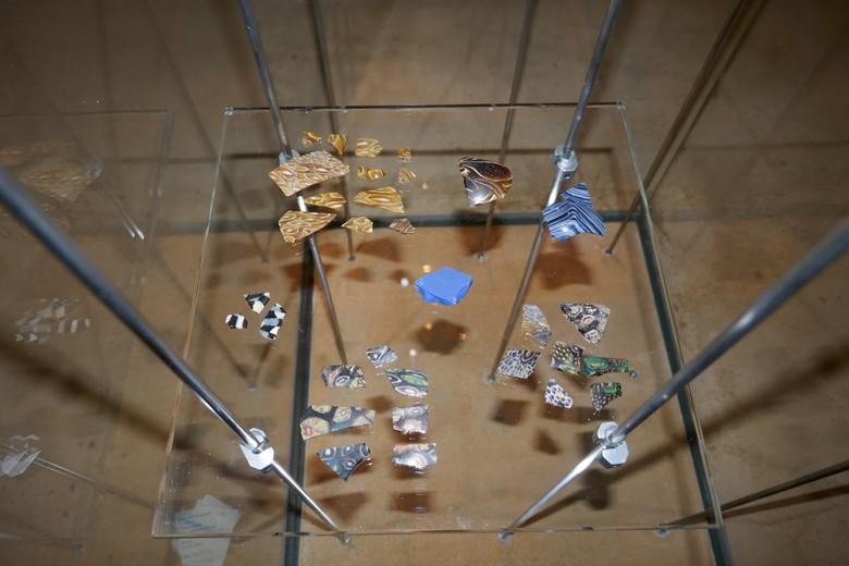 Auf diesem Bild sind die Millefioriglasscherben zu sehen. Sie liegen in einer Vitrine und sind dort in verschiedenen Gruppen nach Muster und Farbe sortiert. Oben links im Bild liegt eine Gruppe mit 12 gelben Scherben, die ein augenähnliches Muster in weiß aufzeigen. Rechts daneben liegt eine einzelne Scherbe in braun mit weißen Fäden als Muster. Daneben liegen, am rechten Bildrand, zwei blaue Scherben, die mit weißen und hellblauen Fäden durchzogen sind. In der Mitte liegt eine blaue undurchsichtige Scherbe ohne Muster. Links am Bildrand sind drei Scherben mit einem weiß, schwarz und blau kariertem Muster. Im unteren Bereich des Bildes sind zwei Scherbengruppen, die Scherben mit bunten Augen- und Blumenmustern enthalten. Es sind insgesamt 33 Scherben. Das Foto wurden von schrägoben geschossen.