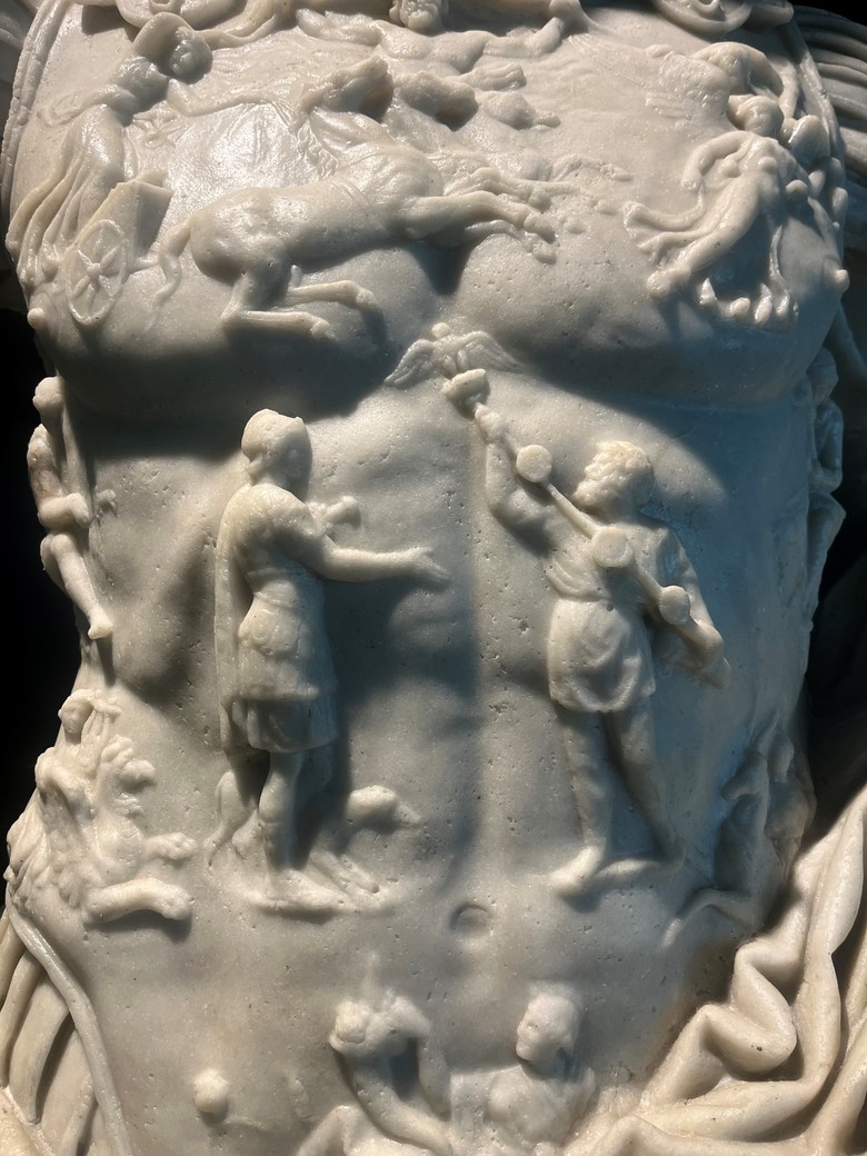 Das Bild zeigt eine Nahaufnahme des Brustpanzers der Augustusstatue. In der Mitte stehen sich zwei Figuren gegenüber. Die linke Figur trägt ein kurzes Gewand und streckt den rechten Arm nach vorne aus. Die rechte Figur trägt lange Kleidung und hält mit erhobenem Arm einen Stab mit mehreren runden Scheiben, am oberen Ende thront ein Vogel. Die rechte Figur scheint den Stab an die linke zu überreichen. Oberhalb dieser Szene, auf der Brust, sind weitere Figuren zu erkennen, darunter eine Figur auf einem Wagen mit zwei Rädern, vor dem Pferde gespannt sind sowie eine liegende, menschliche Gestalt. Entlang der Ränder befinden sich noch weitere Figuren und Ornamente, welche die mittlere Szenerie umrahmen.