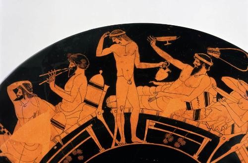 Zu sehen ist die eine Seite der Außenseite der Kylix. Auf dem Bildausschnitt sind vier Teilnehmer des Symposions zu erkennen. Alle Teilnehmer tragen einen Kranz um den Kopf. Drei bärtige, unbekleidete Teilnehmer liegen auf Speisesofas. Der linke von ihnen nimmt eine anmutige Pose ein, bei der er einen Arm an den Kopf legt. Der zweite von ihnen spielt eine Doppelflöte. Der dritte von ihnen schleudert eine Kylix am Finger. In der Mitte der Szene, zwischen den Speisesofas, steht ein bärtiger Teilnehmer mit langen Haaren, der einen Krug hält und Wein nachschenken möchte. Vor den Speisesofas befinden sich niedrige Tische, an denen Efeublätter hängen. Rechts oben schwebt über dem Kopf des dritten, bärtigen Teilnehmers eine Leier.