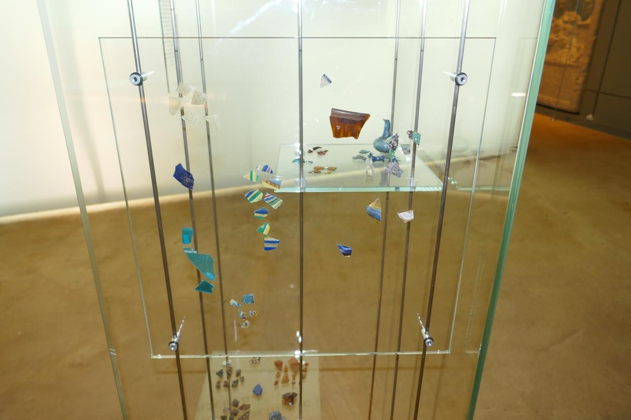 Zu sehen sind römische Glasscherben aus Haltern in einer Vitrine. Die Vitrine ist mit vielen bunten Scherben in verschiedenen Farben ausgestattet, die sowohl hängen als auch liegen.