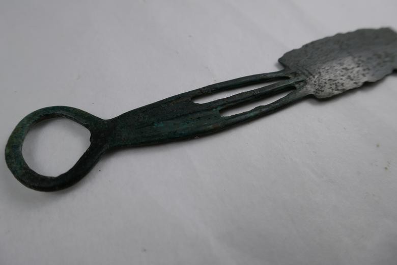 Nahaufnahme des dreigeteilten Griffs mit geschlossenem Endring eines zweischneidigen Bronzerasiermessers aus der späten Bronzezeit. Die drei durchbrochenen Stege sind gut erkennbar. Fotografiert auf hellem Untergrund, leicht seitlich von schräg unten.