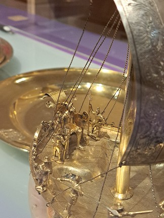 Zu sehen ist eine Nahaufnahme der Person auf dem Thron, der auf dem Tafelschiff am Heck auf einer leicht erhöhten Plattform steht. Der Thron ist mittig im Bild zu sehen. Alles ist golden. Die Lehne des Throns ist nach hinten gebogen und das Ende bildet eine Rolle. Außerdem kommen aus der Lehne an jeder Seite ein Kringel. Die Armlehnen sind weit und haben am Ende auch einen Kringel. Hinter dem Thron, am Ende des Schiffs, sind wieder Verzierungen. Auf dem Thron sitzt ein Mann, der einen Hut trägt, der aussieht wie ein Hexenhut. Er trägt Kleidung wie man sie im 17. Jahrhundert getragen hat. In seiner rechten Hand hält er ein Zepter. Der Mann reckt sein Kinn in die Höhe. Neben dem Thron steht jeweils ein Soldat mit Helm und Lanze.  Links am Rand, auch beim Thron, steht ein Männchen mit Hut und Gewehr und vor ihm steht ein Soldat, der eine lange Lanze waagerecht nach rechts hält. Die Spitze zeigt also fast auf den Betrachter, aber die beiden Männchen und die Lanze sind nur unscharf zu sehen. Die Person auf dem Thron ist etwas größer als die anderen, aber alle sind vergoldet. Bei der Erhöhung, auf der der Thron steht, sieht man , dass das Gold schon abgegangen ist, denn es kommen silberne Stellen zum Vorschein. Der Anfang des Masts des Schiffs ist rechts im Bild. Rechts im Bild ist auch ein verzierter Streifen des silbernen Segels verschwommen zu sehen. Viele feine, silberne Ketten verlaufen durch das Foto und sind dann am Schiffsrand fest gemacht. Die meisten der Ketten auf diesem Foto verlaufen nach hinten und sind neben dem Thron befestigt. Unter dem Schiffsdeck lugt links noch ein Stückchen rosa Nautilusgehäuse hervor.  Im Hintergrund ist ein großer goldener Teller zu sehen, der teilweise vom Tafelschiff überdeckt wird und durch die Lampen über der Vitrine leuchtet. Der Untergrund auf dem die Objekte stehen ist ein hellblaues Glas. Oben im Hintergrund sieht man das Vitrinenglas und dahinter einen dicken lila Rand, der verschwommen ist. Die braune Wand hinter der Vitri