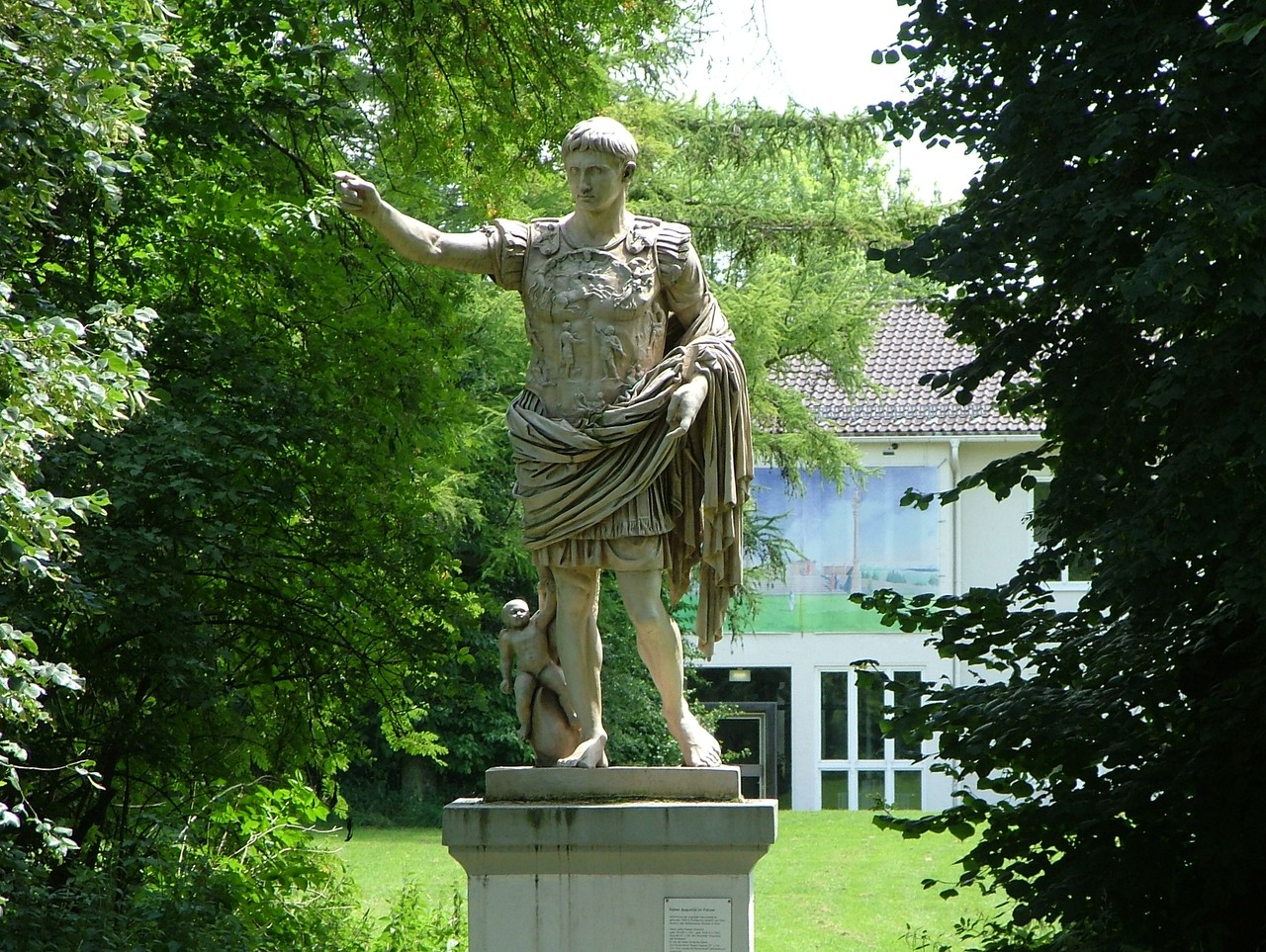 Das Bild zeigt eine farblose, römische und überlebensgroße Statue von Kaiser Augustus auf einem Steinsockel in einer parkähnlichen Umgebung mit dicht beieinander stehenden, grünen Bäumen. Augustus ist als junger Mann, mit einem entschlossenen Blick, dargestellt. Er trägt einen reich verzierten Brustpanzer mit Reliefdarstellungen, einen kurzen Gewandrock und darüber einen Mantel, dessen Enden über seinem linken Arm hängen. Sein rechter Arm ist nach vorne gestreckt und er ist barfuß. Unten seinem rechten Bein befindet sich eine kleine Kinderfigur, die auf etwas sitzt, dass man nicht ganz genau erkennen kann.