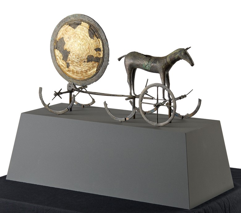 Das Foto eines bronzenen Pferdes auf Rädern, das eine Bronzescheibe hinter sich herzieht. Es steht auf einem grauen Podest, welcher auf einem schwarzen Tuch steht. Die Bronzescheibe ist mit abblätternder Goldfolie verziert und steht ebenfalls auf Rädern. Das rechte Vorderrad ist noch intakt, während von den restlichen Rädern des Pferdes und der Scheibe nur noch die Hälfte vorhanden ist.