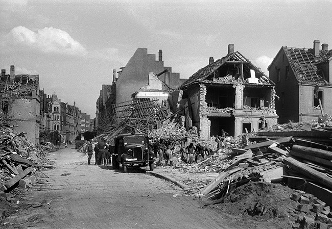 Foto von einer weitgehend Zerstörten Straße in Münster nach einem Bombenangriff Anfang Juli 1941. Die Häuser im Vordergrund sind komplett vernichtet. Die Häuser weiter hinten stehen zwar weites gehend noch sind aber nicht mehr bewohnbar. Man kann einige Menschen in der Uniform der Nationalsozialisten an einem Auto stehend neben den zerstörten Häusern sehen.