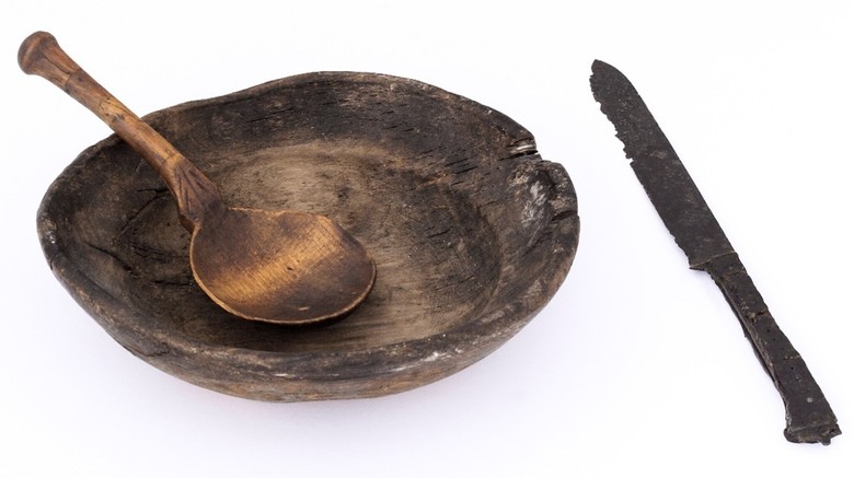 Ein hölzerner Teller auf weißem Untergrund, in dem Teller liegt ein hölzerner Löffel und rechts daneben ein altes Messer.