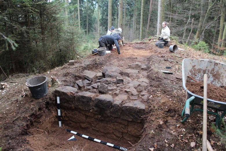 Vier Archäolog:innen arbeiten in warmer Kleidung umgeben von Eimern und Werkzeugen an der Ausgrabung der gemauerten Esse der Schmiede. Im Hintergrund ist ein Wald zu sehen.