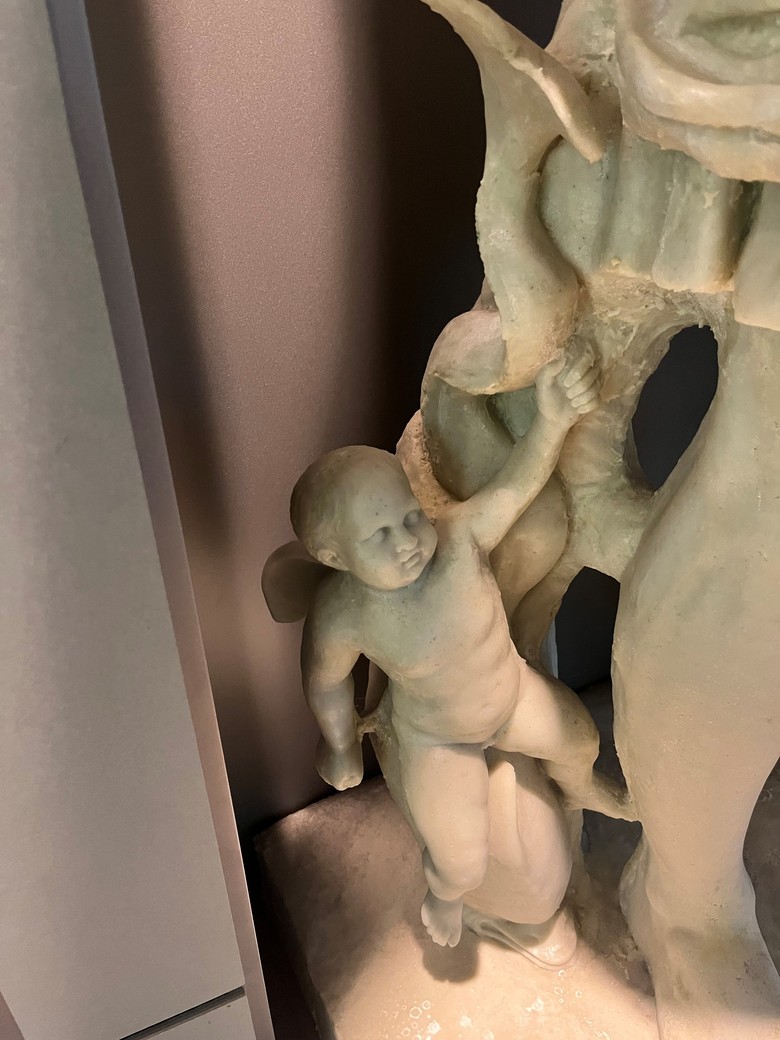 Auf dem Bild ist die nackte Figur eines kleinen Kindes zu erkennen, die auf dem Rücken mindestens einen kleinen Flügel trägt und auf einem Delfin mit geschlängelter Flosse sitzt, der nach unten gerichtet ist. Diese Figuren befinden sich an dem rechten Bein der Augustusstatue.